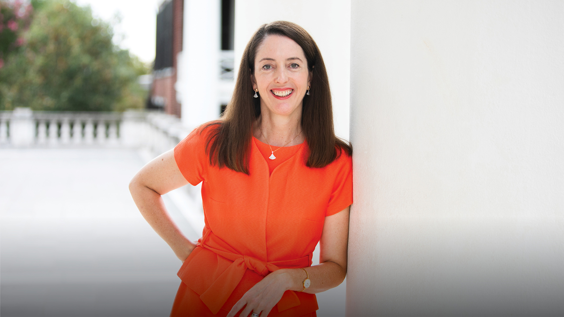 Woman with long brown hair wearing orange dress leans against the outside corner of a white building and smiles.
